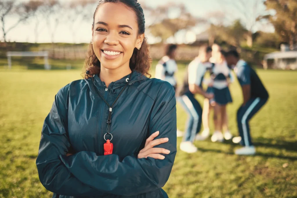 Treinadora sorrindo em frente ao campo com a turma ao fundo